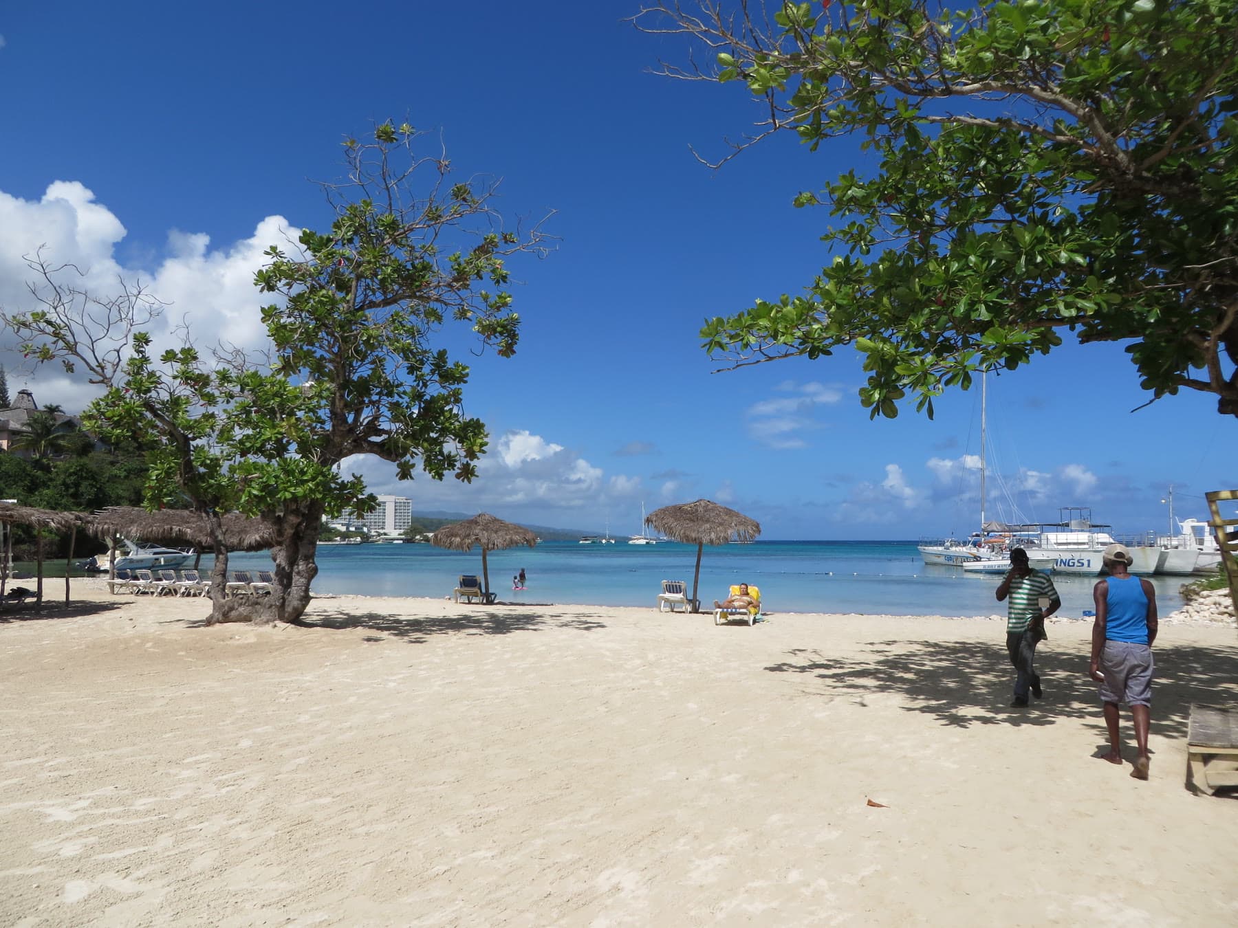 Mahogany Beach in Ocho Rios, with lounge chairs, thatched umbrellas, boats, and a hotel along the bay